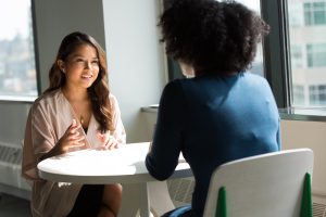 Two women sit across from each other at a table in an office setting.