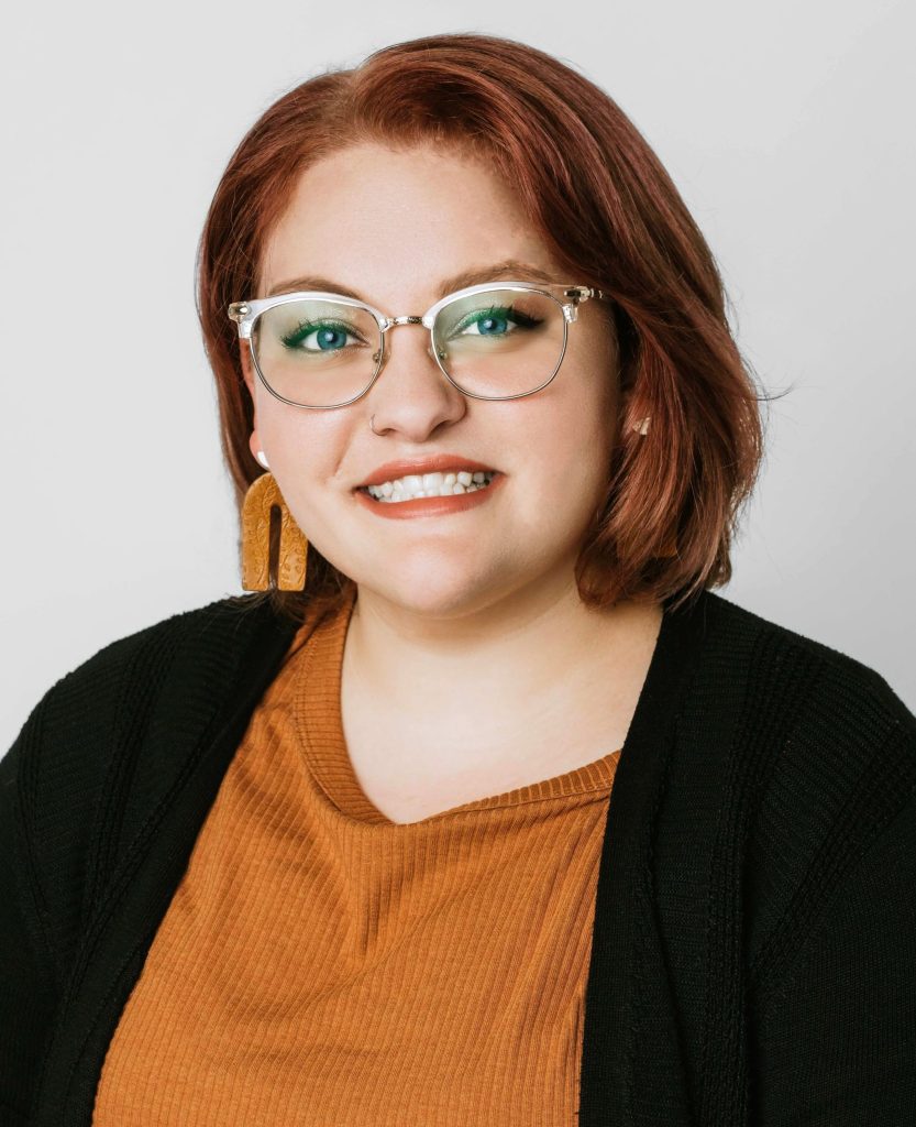 Corinne Farleigh has red hair and is smiling, wearing gold-rimmed glasses, orange earrings, and an orange shirt.