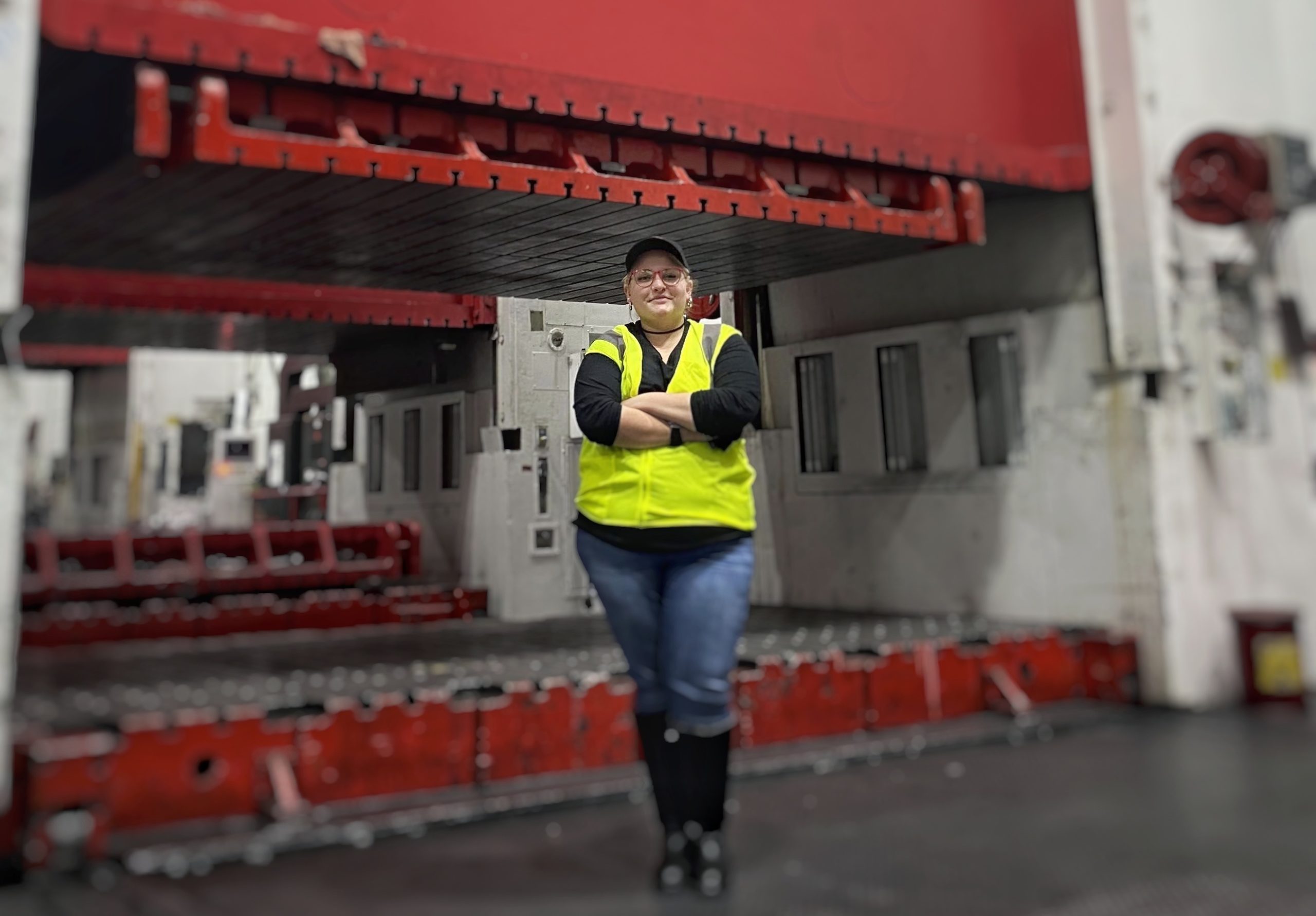 Corinne Farleigh is standing confidently wearing a safety vest with red industrial equipment behind her.
