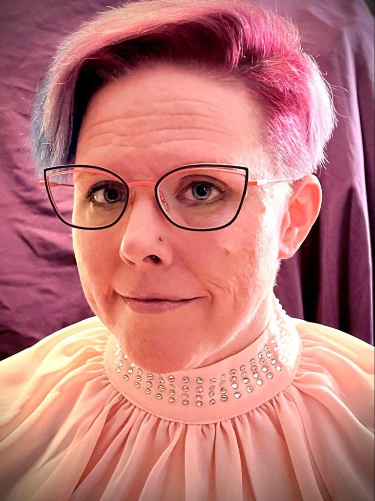 Headshot of Cynthia Hurlbert who is wearing cat-eye-shaped glasses and has short pink hair.