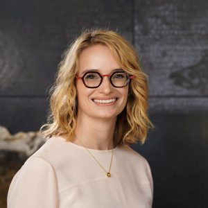 blond woman wearing glasses (black frames with red arms and nose bridge) and light pink shirt smiles for the camera