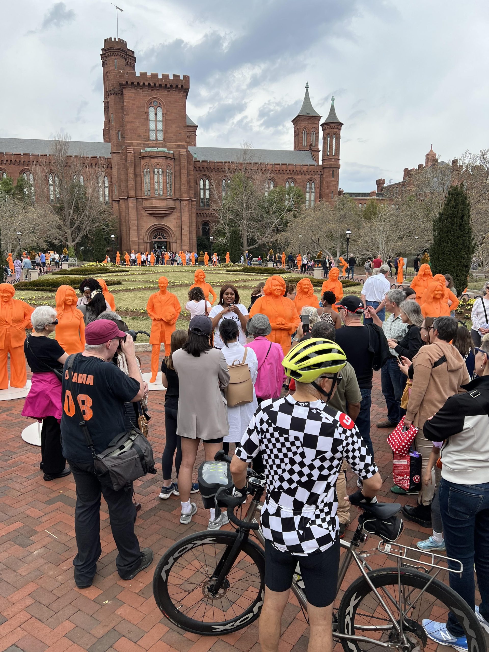 Dr. Lataisia Jones is speaking next to her statue to a large crowd of people. The Smithsonian castle, garden, and more orange statues are in the background.