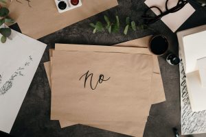 A stack of brown paper with "no" in cursive handwriting surrounded by aesthetically pleasing items on a desk, like plants, glasses, and a paint pallette