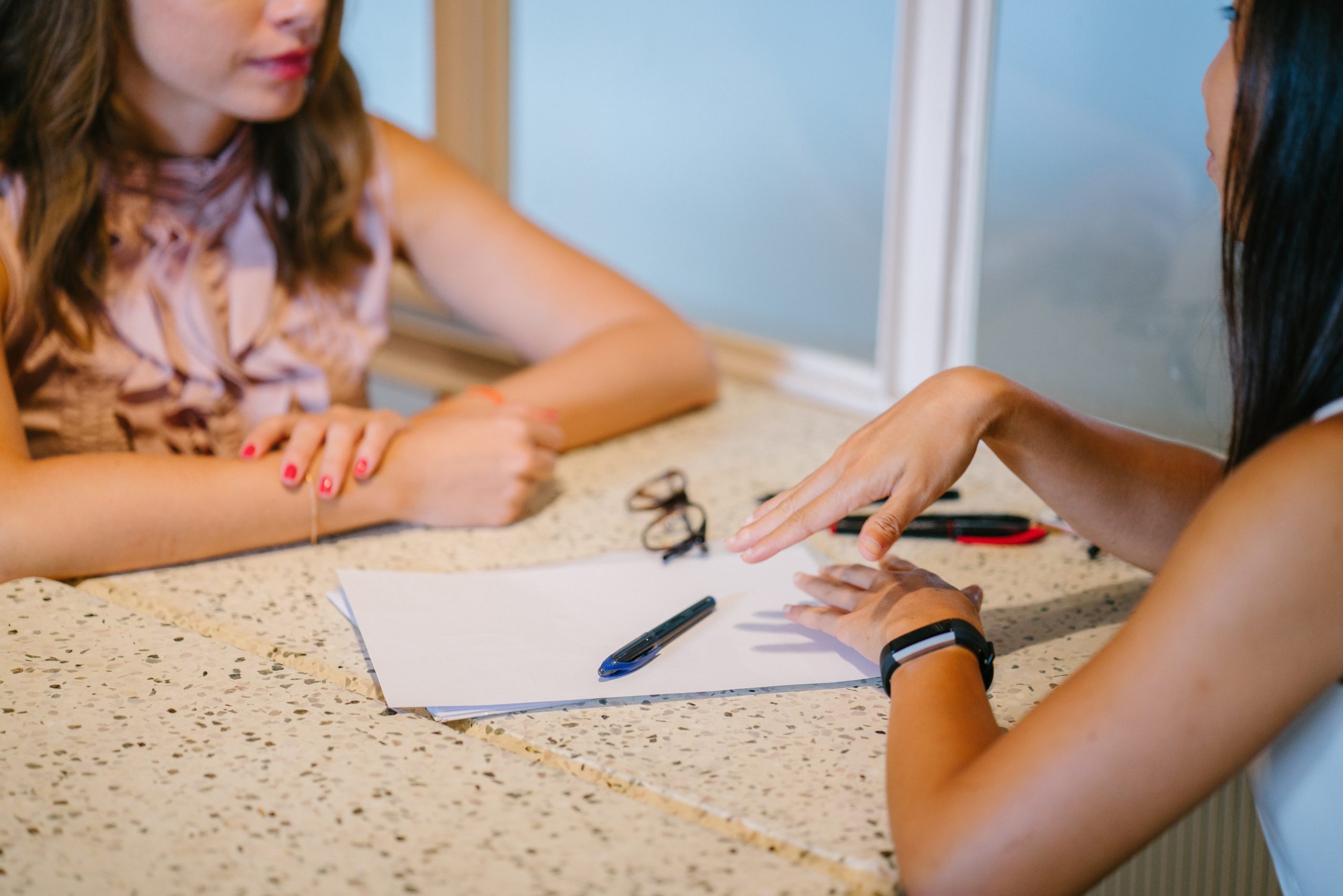 Two people sit across from one another with a paper and pens on the table between them