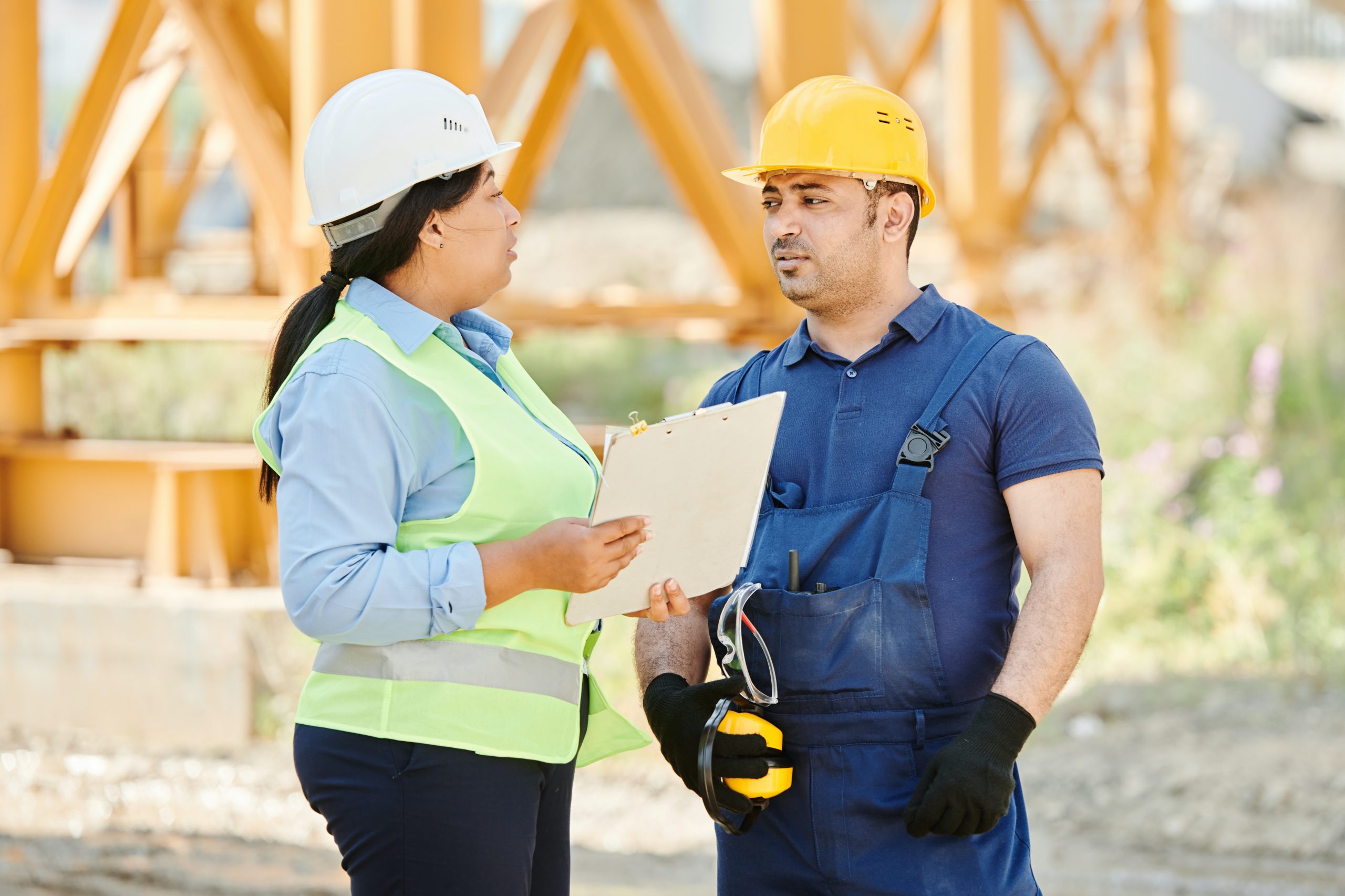 A woman wearing a hard hat and safety vest holding a file folder speaks to a man wearing a hard hat, coveralls, and work gloves with a steel structure behind them.
