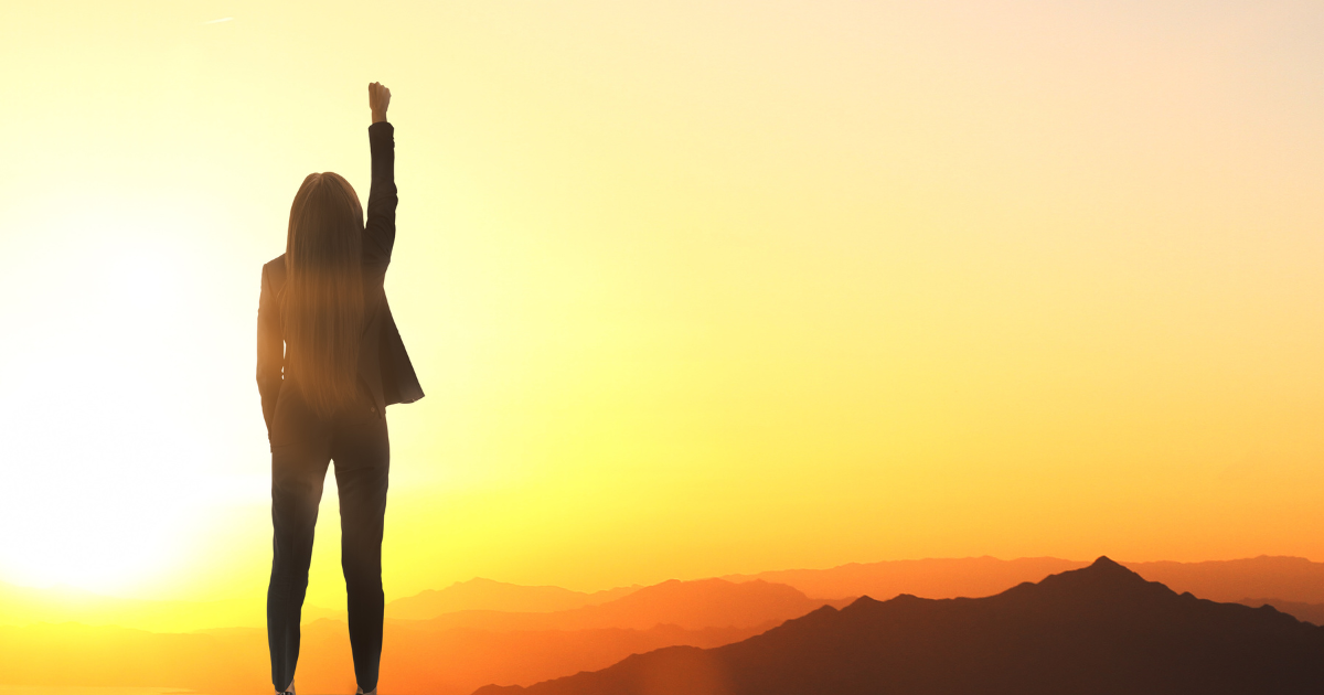 A woman stands on a mountain at sunset (or sunrise) with her back to the camera and a fist in the air