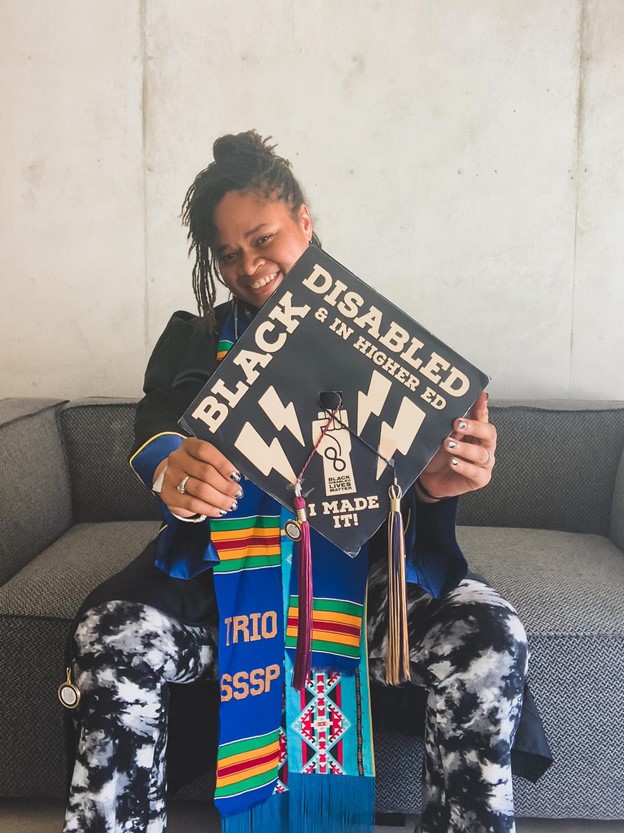 Syreeta Nolan is smiling and wearing her graduation gown. She is holding a graduation cap in front of her that's decorated with the words "Black, Disabled in Higher Ed. I made it!" and a Black Lives Matter symbol.
