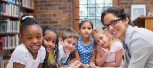 Woman teacher wearing glasses leans on a table surrounded by several elementary students