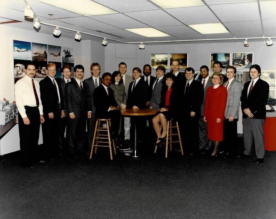 A group of several men and 3 women in an office in the 80s